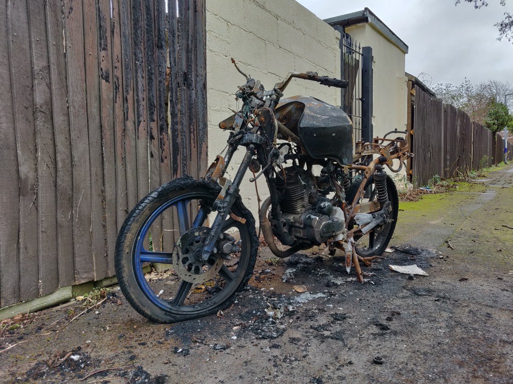 an abandoned motorbike that has been stripped by fire, leaving only the metalwork of the frame, the engine and the rubber on the tyres 