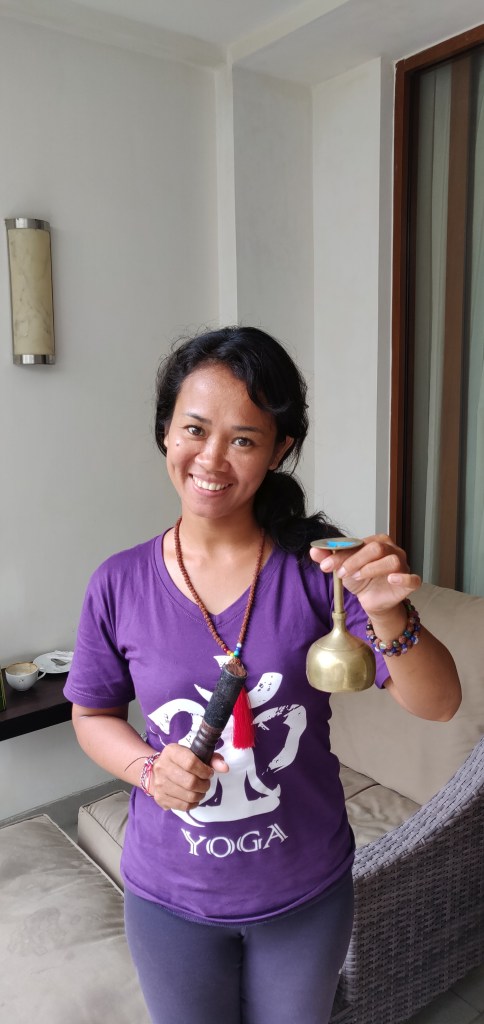 Komang Sukerni wearing a purple t-shirt with a white yoga print standing in front of wicker rattan furniture and holding a tiny singing goblet