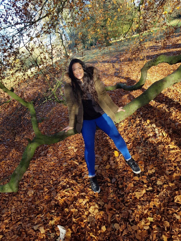 Beautiful smiling Rooby with hair down wearing blue jeans and an olive green coloured winter jacket sitting on a branch above autumn leaves  
