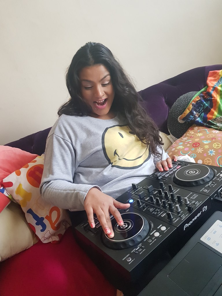 Rooby wearing a grey sweatshirt with a yellow smiley face design looking excited sitting on a sofa with a Pioneer DJ music mixing device on her lap