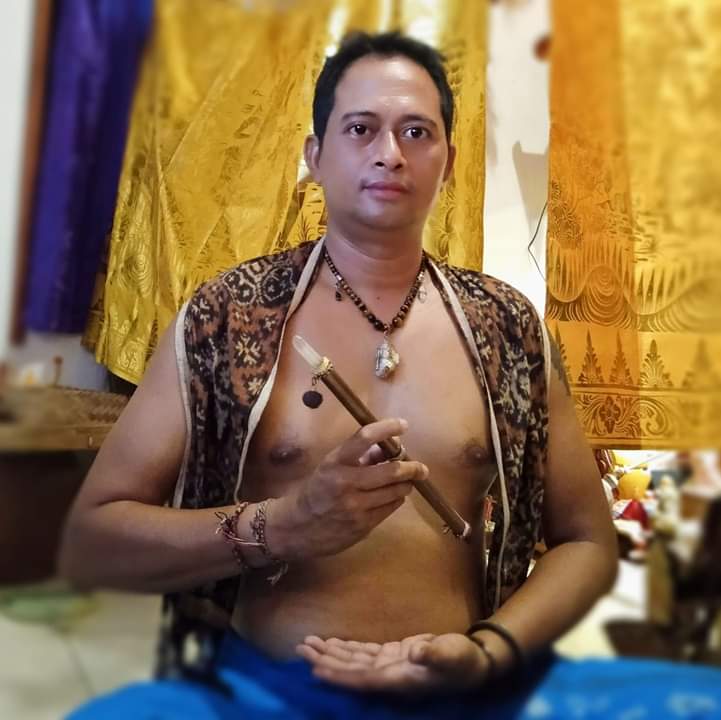 Agung Putrawan sits cross legged wearing a vest patterned with different shades of brown and holding ceremonial paraphenalia 