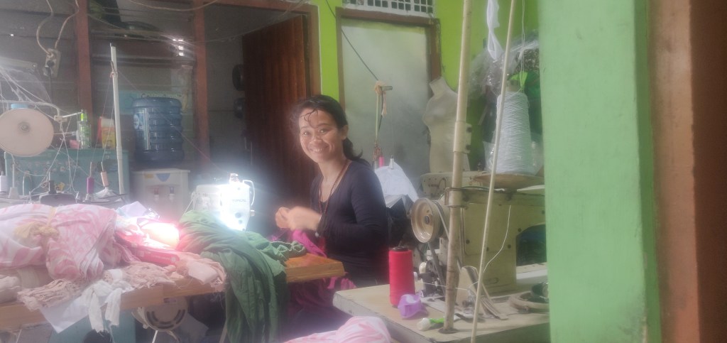 A smiling Komang Sukerni sitting at a sewing machine illuminated by the sun and surrounded by fabric and yarn in a room with green walls   
