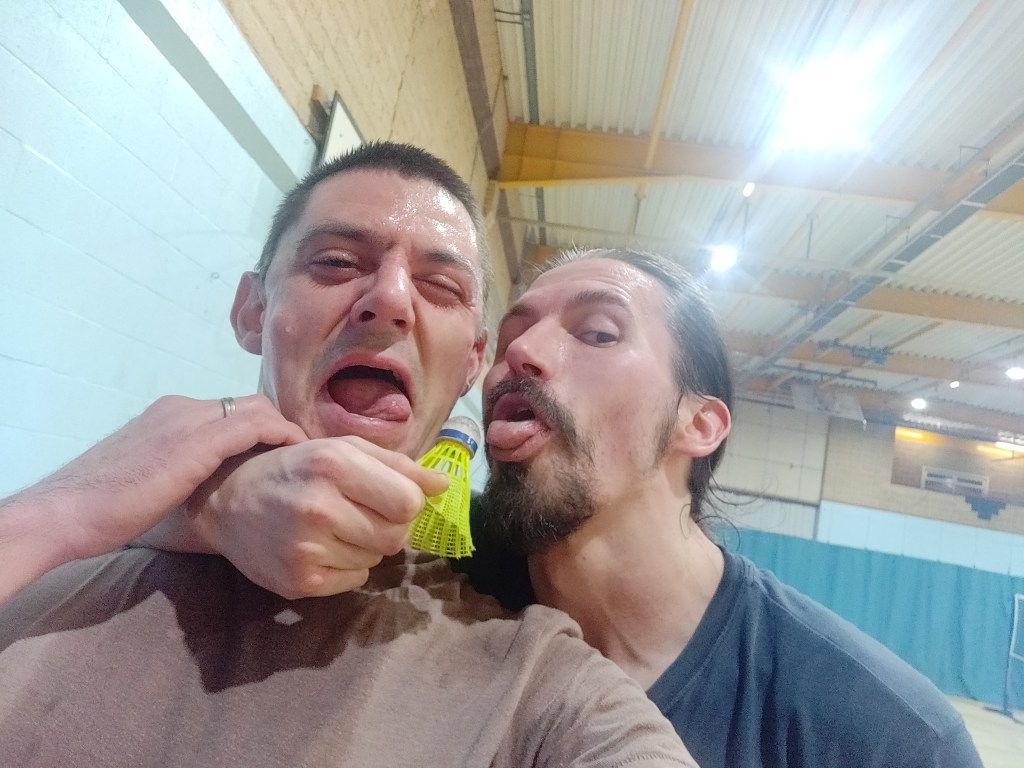 Selfie of James of cafe bistro fame with one arm around Guerrilla Nature and in a sports hall with bright lights above as they both pretend to lick a shuttlecock