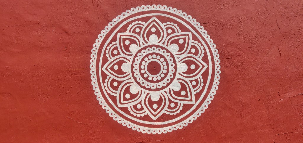 A rendered wall painted a shade of red reminiscent of clay with a circular mandala featuring petal and circle shapes all lit by summer sunshine