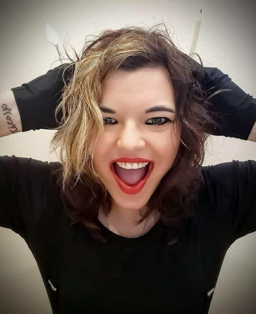 Kirsty Xenia with dark hair on her right side and light hair left with her hands in black gloves behind her head holding a pencil in left hand and scraper in right hand