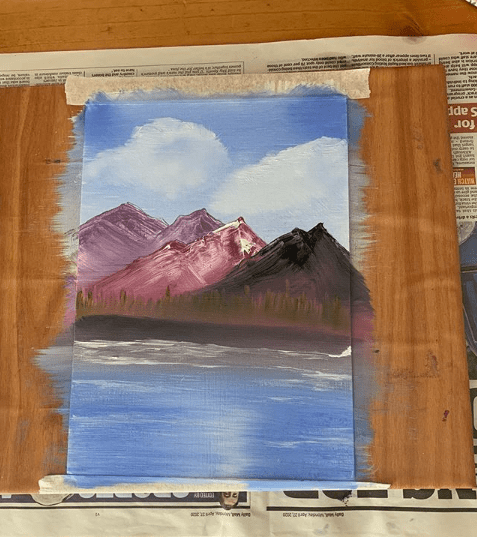 a painting showing a mountain range with fluffy white clouds behind and a tree lined lake in the foreground left to dry on a wooden board