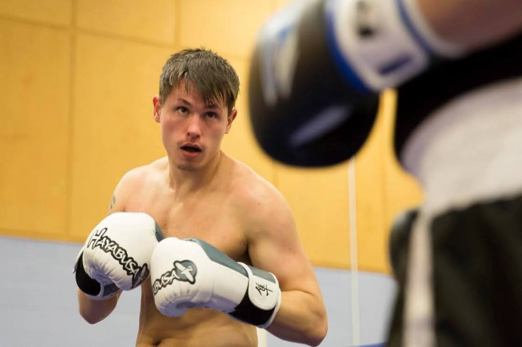 a topless man wearing boxing gloves in focus at the back of the frame looks towards the edge of a waistline and part of a glove in the foreground 
