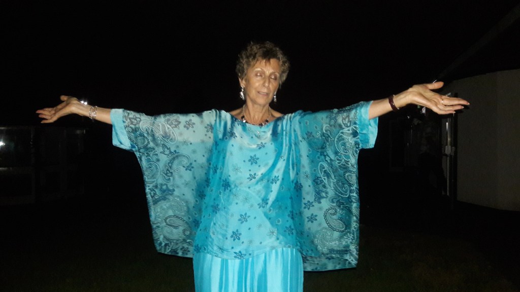 Mary wearing a bright blue dress and a matching shawl covered with intricate patterns stands with her arms outstretched and her eyes closed  