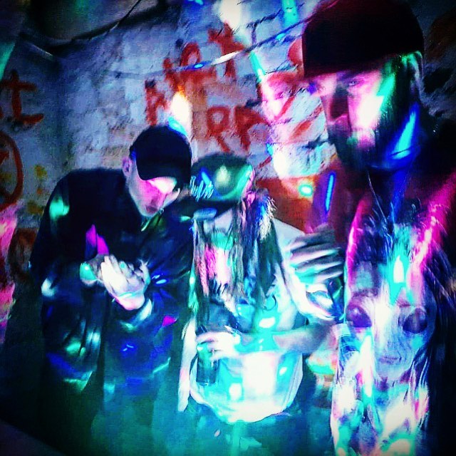 One Dread wearing a baseball cap with two other people in front of a white painted brick wall with graffiti on and multicoloured lighting creating patterns