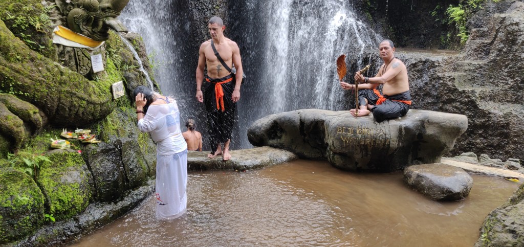Guerrilla Nature stands and Agung Putrawan sits on a big flat rock in front of a waterfall  whilst a woman dressed in white faces away front left
