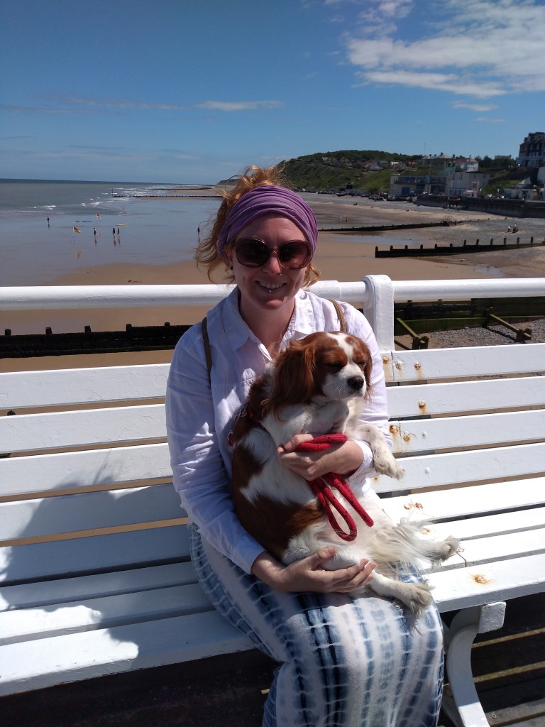 Samsdabblers sits on a white bench in the sunshine on a pier with Iris the dog on her lap both facing front with blue sky and the beach in the background 