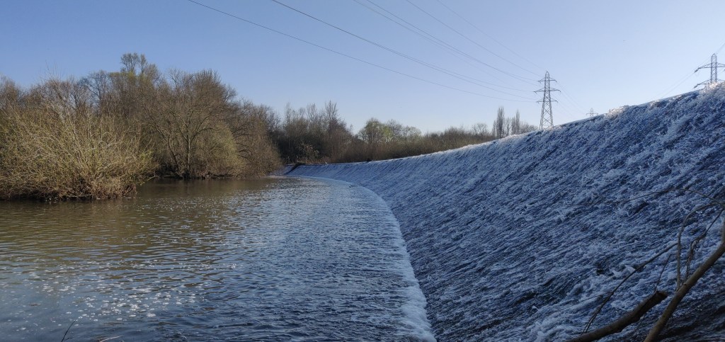 A long weir stretches from bottom right corner to left of centre and is lined by trees to the left and electricity pylons to the right of the azure sky background