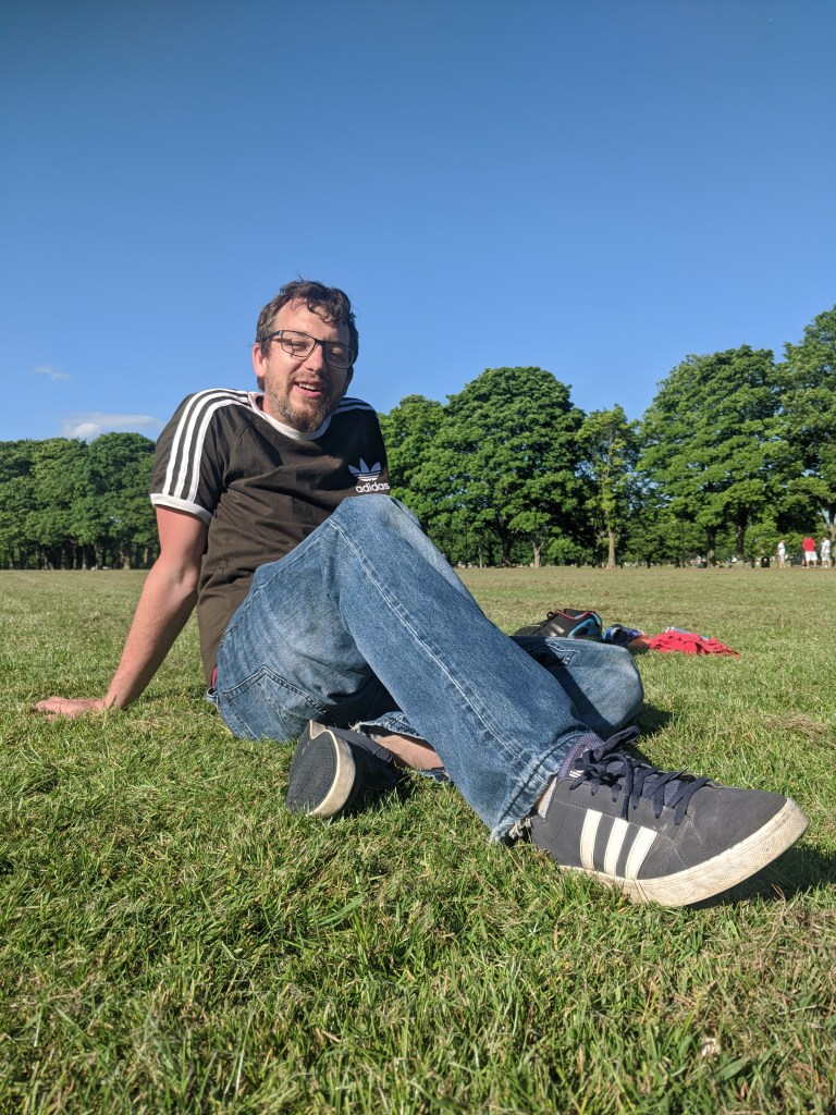 low angle shot of a smiling man named Hodge sitting wearing glasses and a t-shirt and on the grass in the summer sunshine with a clear blue sky 