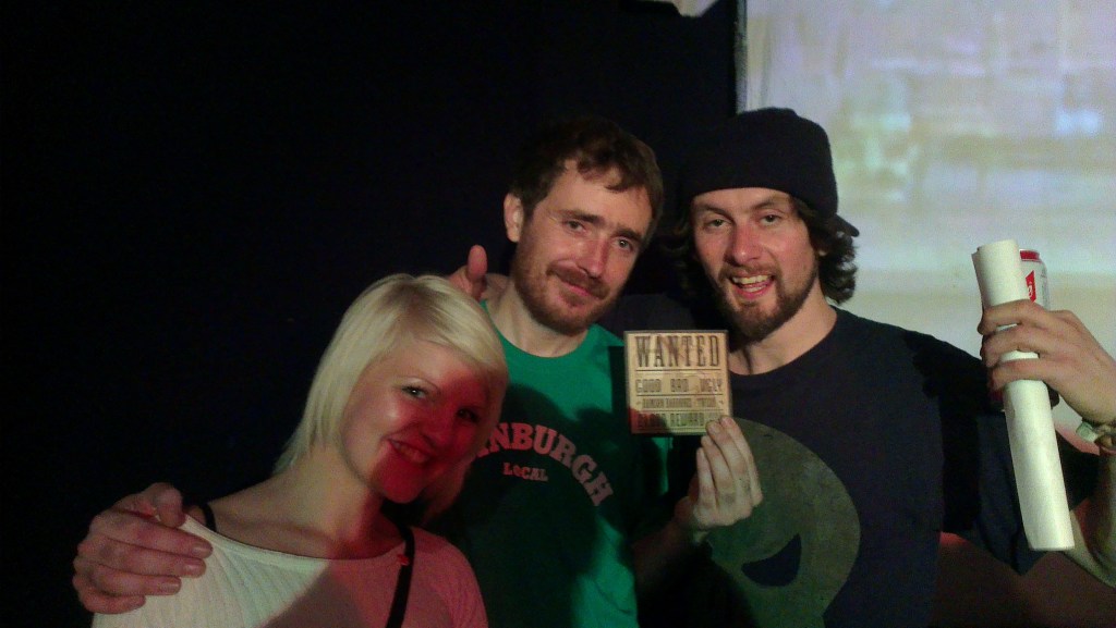 Chester P holds up a copy of Wanted by Ransom Badbonez in his left hand and his right hand on a woman's shoulder with a man slightly behind to the right