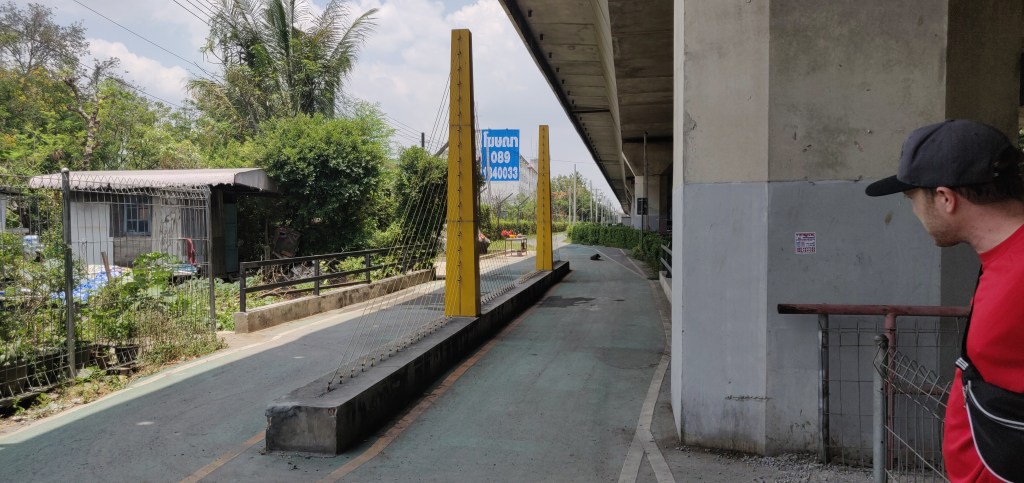A caucasian man stands to the far right below a raised raised road, looking away into the central distance along another toad towards a weird structure 