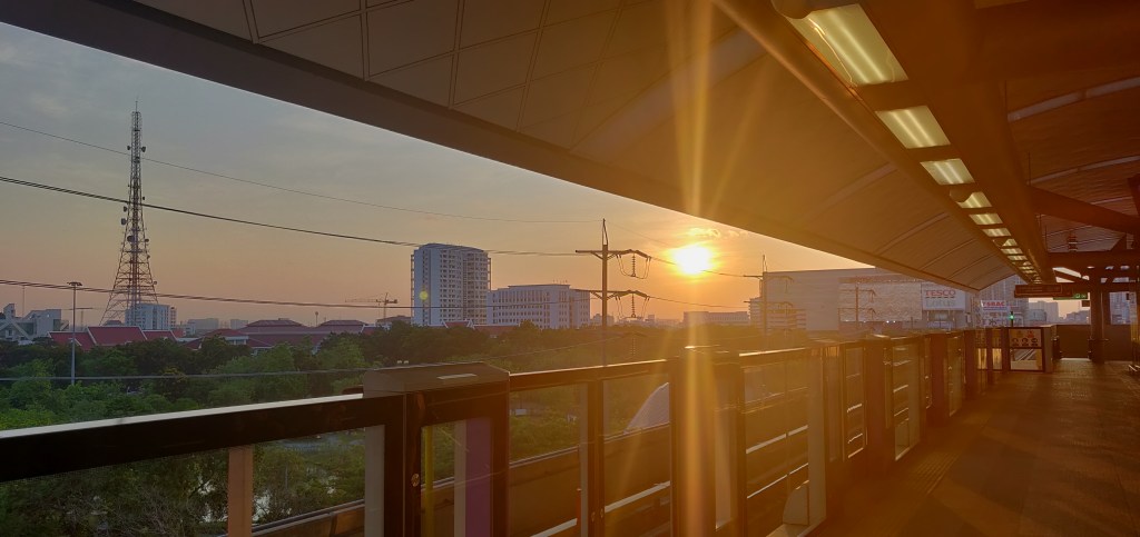 The early morning sun rises behind clouds in the distance, illuminating a city scape showing high rise buildings in the distance and a train platform close 