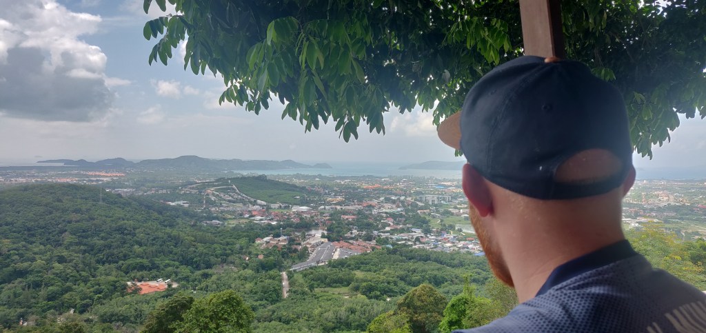 The back of a caucasian man's head sporting a dark blue baseball cap looks out over the distant Bay of Chalong from Phuket View Cafe in Thailand