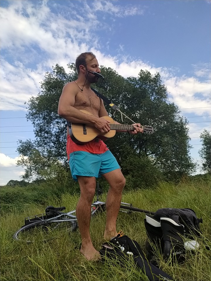 Ben Taylor with bare feet and wearing shorts stands on grass playing a harmonica on a frame and a small acoustic guitar with blue sky behind