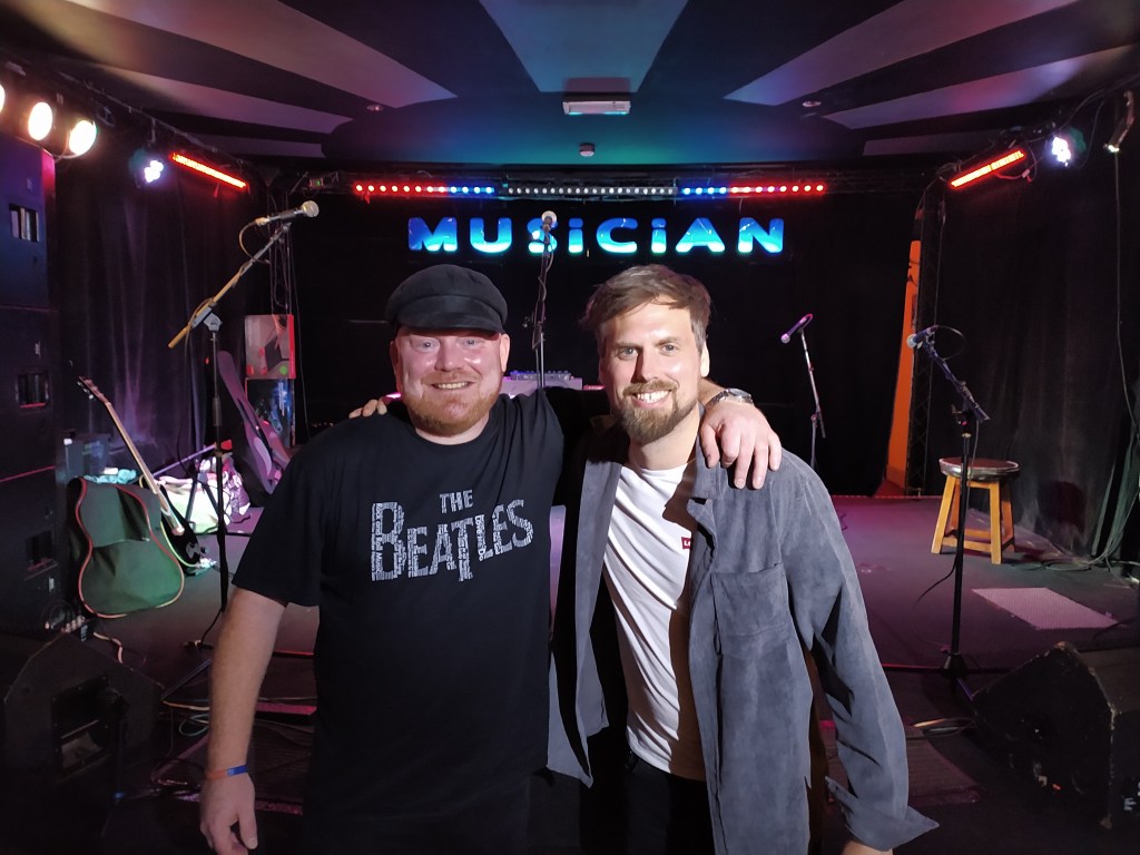 Daz Lynch wearing a Beatles t-shirt and Mark Ryman wearing a white t-shirt under an open blue shirt in front of the stage at The Musician in Leicester