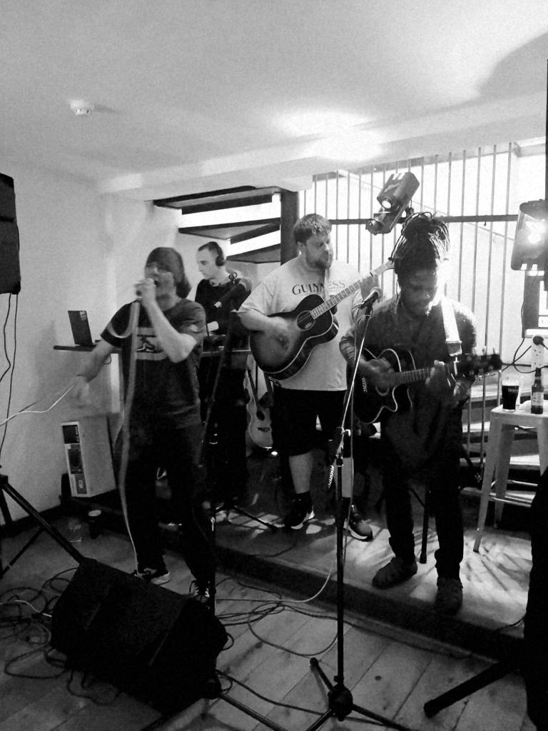 Digital greyscale photograph of Calvin J, Alex Hopps play guitars as Low 3 and Guerrilla Nature also play on a very small stage with a staircase behind   
