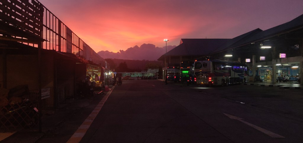 Dusk sky glows red in the distance around where the sun sets behind grey clouds, casting an eerie light on Phuket main bus terminal in the foreground 